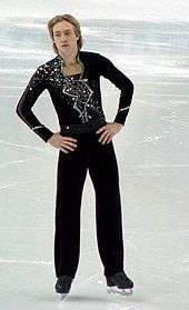 A blond male figure skater dressed in a black suit with glitters moves around on an ice rink.
