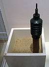 Black club-shaped landmine with a spike on its top end, mounted on the end of a wooden stake which is standing in a sand-filled container.