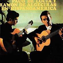 Two men playing flamenco guitar in front of a greenish tinted background depicting a body of water and mountain in the distance.