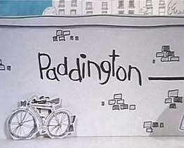 In this animation title card, the name "Paddington" is scrawled on a drawing of a brick wall. A bicycle leans against the wall, and a sliver of blue sky and tall buildings are visible over the wall's top edge.