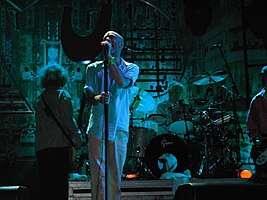 A blue-tinted photograph of musicians performing in front of an industrial background. From left to right: a long-haired male stands with his back to the camera playing bass guitar, a middle-aged Caucasian male sings into a microphone, a middle-aged Caucasian male plays behind a black-and-silver drum set on a riser, and a guitar player is mostly cropped from the extreme left of the photo.