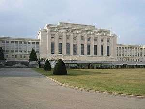 A drive leads past a manicured lawn to large white rectangular building with columns on it facade. Two wings of the building are set back from the middle section.