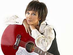 A woman with short brown hair, wearing a white shirt and leaning on a red guitar