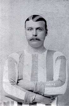 Monochrome photo of head and upper torso of young man with shiny side-parted hair and large moustache, wearing long-sleeved sports shirt with vertical stripes