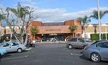 A view of a single-story brown brick building from a parking lot in front, with palm trees on either side of the frame. In the center is a red brick entrance pavilion with "Panorama Mall" written on it in stylized metal letters