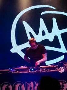 A DJ works with a soundboard before a large Doomtree logo, a crudely drawn crown that has been crossed out