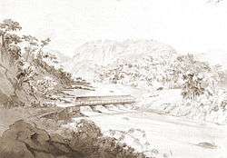 A pencil with wash sketch depicting a forested valley with riders and pack animals on a road on the left side which descends to a covered bridge across a river on the valley floor