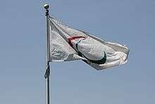 A flag with three red, green, and blue swooshes on a white background. It is attached to a flagpole and is framed by a blue sky