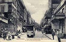 A sepia picture postcard of a commercial street scene looking down the street, showing a tramcar head-on. Some pedestrians mill about.