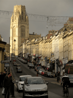 Park Street, Bristol. A steeply-sloped busy shopping street with light stone-coloured Georgian buildings on both sides. At the top is the ornate and dominant tower of the Wills Memorial Building.