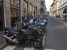 A row of black motorcycles parked in the foreground alongside a narrow street that recedes to near the background. Several men are sitting around and talking on some of the motorcycles, and people walk on the sidewalks on both sides of the street