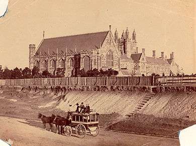 An old sepia photograph shows Sydney University from the west. Only the front section and Great Hall of the Main Building exists. It is separated from the road by a paling fence and a steep eroded incline, where there is now a garden. A horse-drawn bus with passengers up-top is standing in the road.