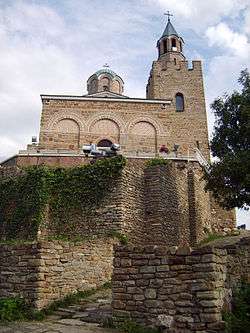 View of a medieval Byzantine-style church's front facade and bell tower slightly from below, with partially destroyed stone walls in the foreground