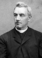The upper-body of a middle-aged male looking slightly to his right. He wears the black robe and white collar of a Catholic priest.