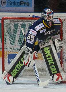 An ice hockey player standing partially crouched in goals. He is wearing a helmet, gloves and leg pads and a blue and yellow uniform.