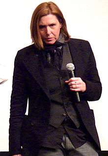 A female musician, Patty Schemel, wearing a black suit and holding a microphone.