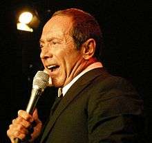 Color bust photo of a man in a tuxedo, holding a microphone.