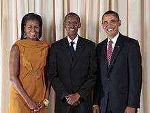 Michelle Obama, Paul Kagame and Barack Obama, standing and smiling in front of a curtain