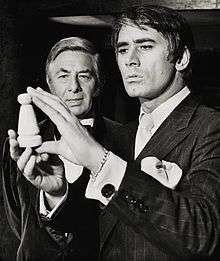 A black-and-white photograph of two well-dressed men examining a small wooden object.