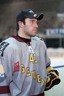 An ice hockey player facing to the right of the camera. He is wearing a baseball style cap and a grey uniform.