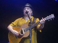 Paul Simon sings live with a guitar in hand.