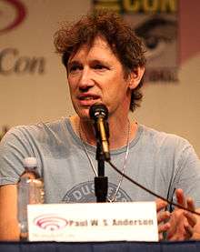 Paul W. S. Anderson, a Caucasian man in his mid-forties with messy brown hair seated in front of a microphone and looking to the side
