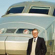 Cropped colour photograph of man in dark jacket, shirt and tie standing in front of French TGV high-speed train.