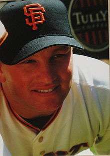 A smiling man wearing a white baseball jersey and black baseball cap with an interlocking "SF" in orange on the face