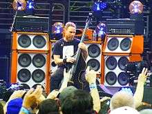 A male bassist, Jeff Ament, playing upright bass in a concert. He is seated in front of several large, tall speaker cabinets.
