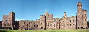 A sandstone castle-like building with a tower slightly to the left of centre. The walls are battlemented and in the foreground is a grassed area