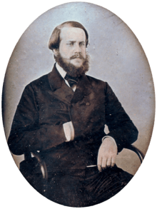 Photographic half-length portrait of a seated bearded man dressed in a dark, double-breasted coat with his right hand tucked inside the front
