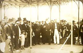 The white-bearded emperor stands among a group wearing formal and military attire under a pavilion.