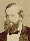 Sepia photograph showing the head and shoulders of a bearded, light-haired man wearing a formal black coat, white shirt and dark cravat