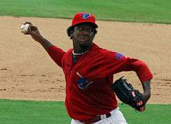 A dark-skinned man in a red baseball uniform and cap throwing a baseball with his right hand