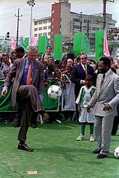 Two men in tailored suits laugh as they juggle a soccer ball between them, watched by a large crowd.