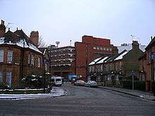A tall red brick building towers over streets of two-storey houses. The roofs of the houses and the surrounding pavements are covered in snow.