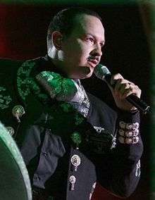 A man dressed in mariachi suit and holding a microphone in his left hand, facing front.