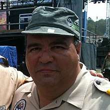 A Hispanic male wearing a beige colored button-up shirt and green cap posing for a picture outdoors.
