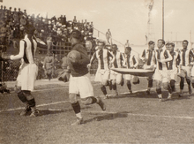 Photo of ten men, running and carrying a large flag, inside a stadium