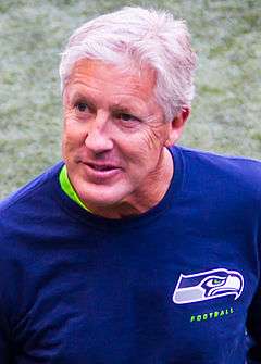 Color head-and-shoulders photograph of silver-haired Pete Carroll in dark blue sport shirt with Seattle Seahawks logo on left breast.