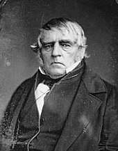 Formal portrait showing the head and shoulders of a white-haired man wearing a dark cape or coat
