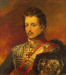 Painting shows a serious-looking young man with a moustache and wavy hair. He wears an elaborate hussar military uniform with gold lace and a gold and red sash over his shoulder.