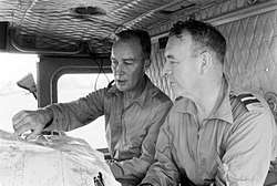 Black and white photograph of two men wearing military uniforms seated in the cabin of a vehicle. The man on the left is pointing at a location on a map.