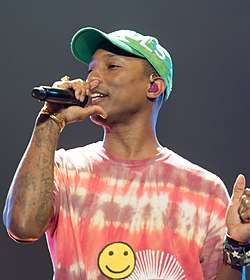 An image of Pharrell Williams holding a microphone in his hand, wearing a red T-shirt along with a green cap.