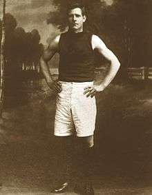 Sepia colours posed photograph of young man standing with hands on hips, wearing a dark sleeveless jumper, long white shorts and black boots