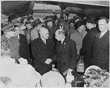 Truman shaking hands with Attlee. A large crowd surrounds them. There is a large propeller-driven airplane in the background.