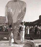 Woman in her thirties in a raincoat and man in his forties dressed in white holding a plastic balloon in front of observers, apparently in a stadium.
