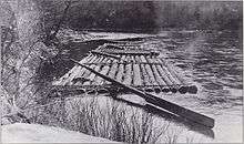 A raft made of long logs lashed together with a large oar for steering is tied to the bank of a stream.