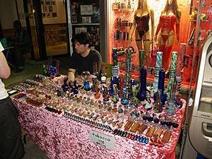 A man sitting at a table on a sidewalk. On the table are an array of pipes from large and ornate to small and plain. A sign taped to the tablecloth in front says "Tobacco use only."