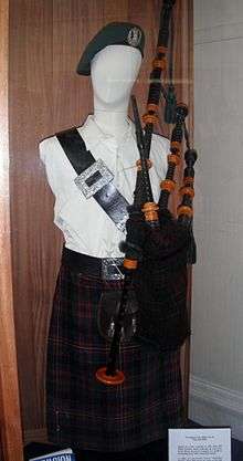 Photograph of Millin's uniform displayed on a mannequin in a museum case, consisting of a black bonnet, a white shirt, a wide black belt with a large silver buckle draped across the chest from the right shoulder to the left side of the waist, bagpipes, and a dark multicolored tartan kilt
